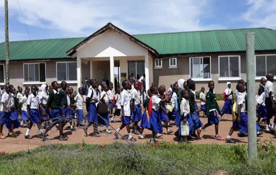Pupils in blue and white uniforms walk in front of a school building with a green roof
